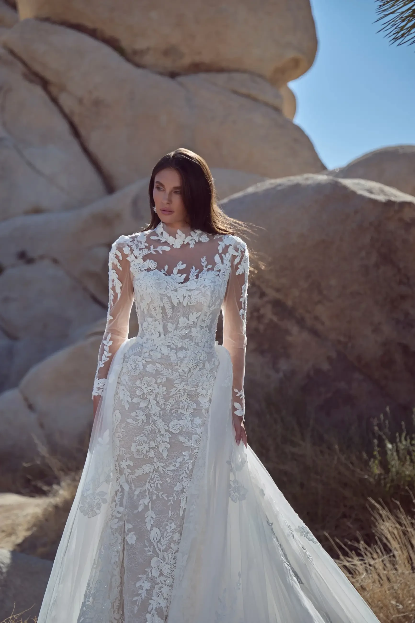 A woman wearing a lace wedding dress with long sleeves, standing among rocky terrain and natural surroundings.