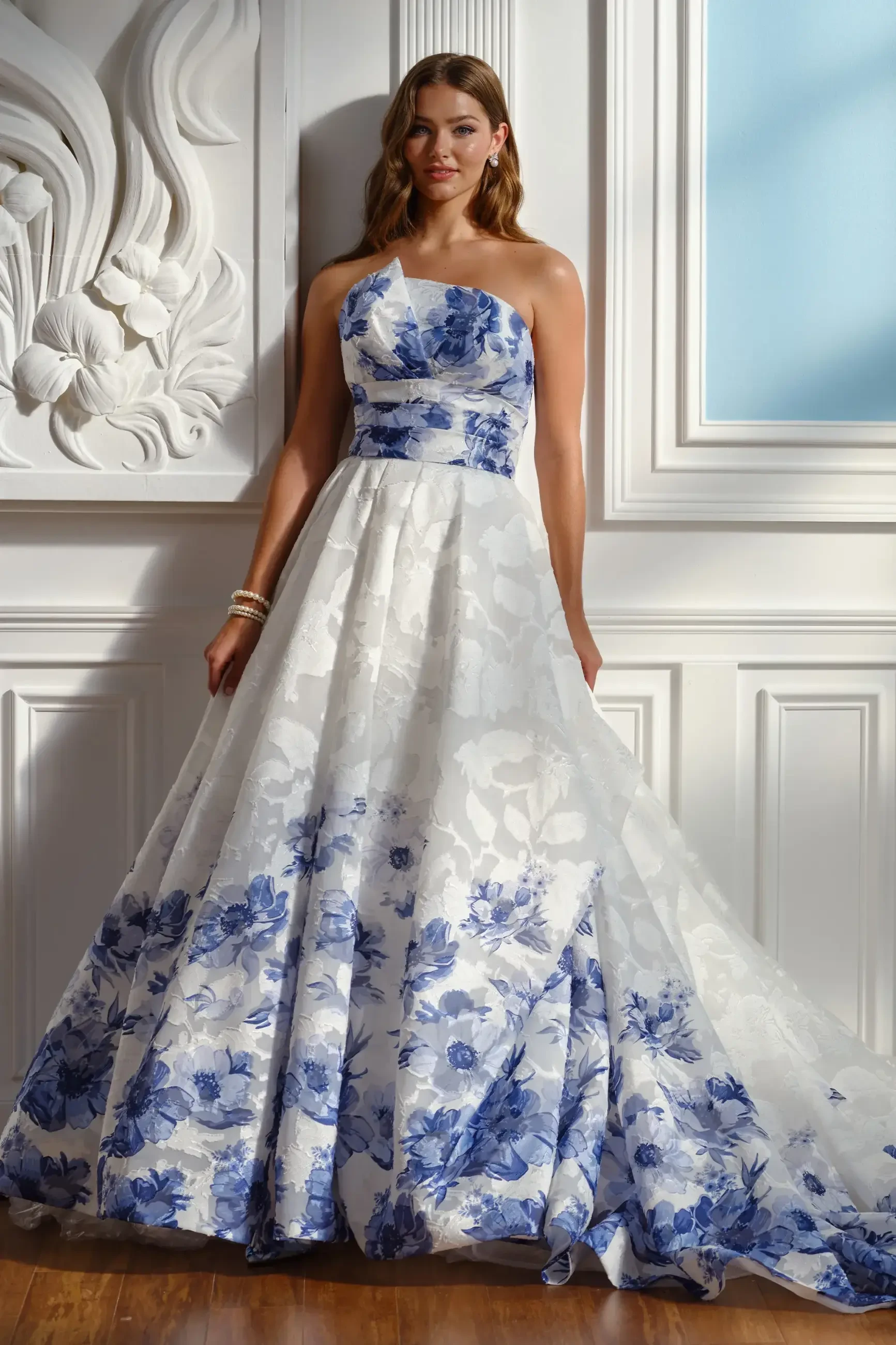 A woman in a strapless, floor-length gown with a floral pattern in blue and white, standing against an ornate white wall.