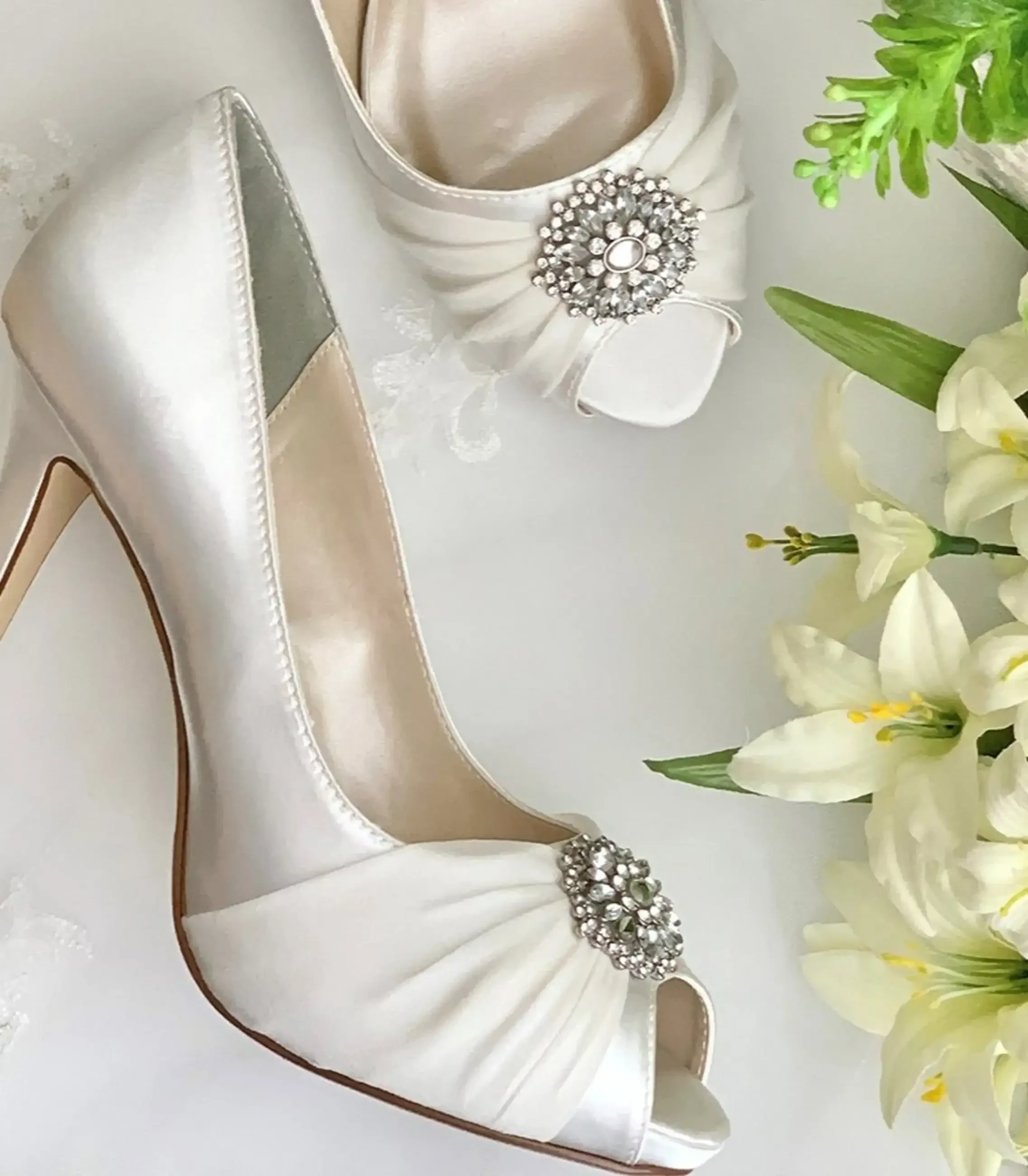 White satin bridal heels with jeweled brooch details beside white flowers.