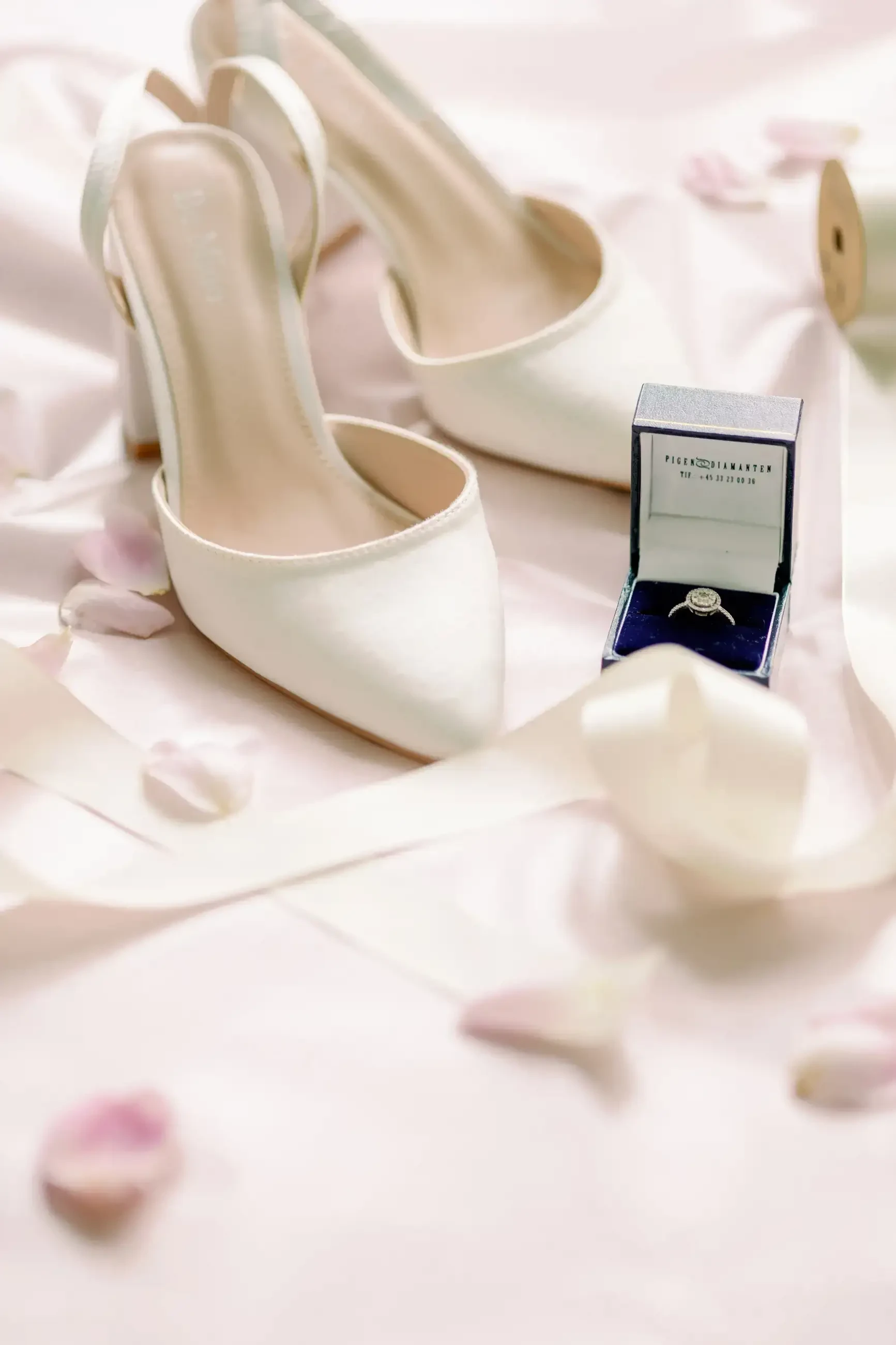 White bridal shoes and engagement ring box arranged on soft pink satin.
